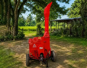 Red mobile wood chipper on a wheeled trailer in a rural yard with trees and a shed nearby.