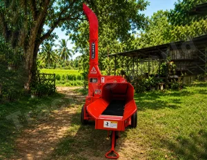 Red wood chipper on a dirt path in a sunny garden, with trees and a shed in the background.