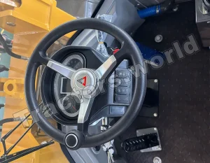 Steering wheel and instrument cluster inside a yellow construction vehicle cab, with control panel to the right and floor pedals below.